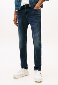 Mężczyzna w ciemnoniebieskich dżinsach, białych trampkach i jeansowej kurtce, stojący na jednolitym białym tle, z jedną ręką w kieszeni.