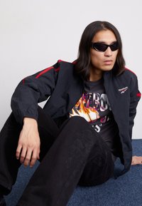 Chaqueta de nailon negra con acentos rojos, camiseta gráfica con llamas coloridas, pantalones negros con bordados florales y gafas de sol.