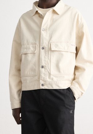Personne portant une veste crème boutonnée avec deux poches à rabats à l'avant et un pantalon noir avec un petit logo Calvin Klein sur la cuisse.