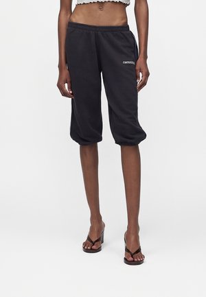 Personne portant un pantalon de survêtement noir raccourci avec des ourlets élastiques et des sandales noires à talons, debout devant un fond blanc uni.