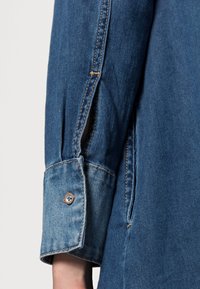 Koszula dżinsowa w jasnoniebieskim kolorze, z mankietem zapinanym na guziki i widocznym przeszyciem. Zawiera boczny rozcięcie dla dodatkowego detalu.