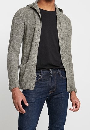 Mann trägt einen grauen, gestrickten Kapuzenpullover mit Reißverschluss über einem schwarzen T-Shirt und dunkelblauen Jeans, steht mit locker neben dem Körper hängenden Armen.