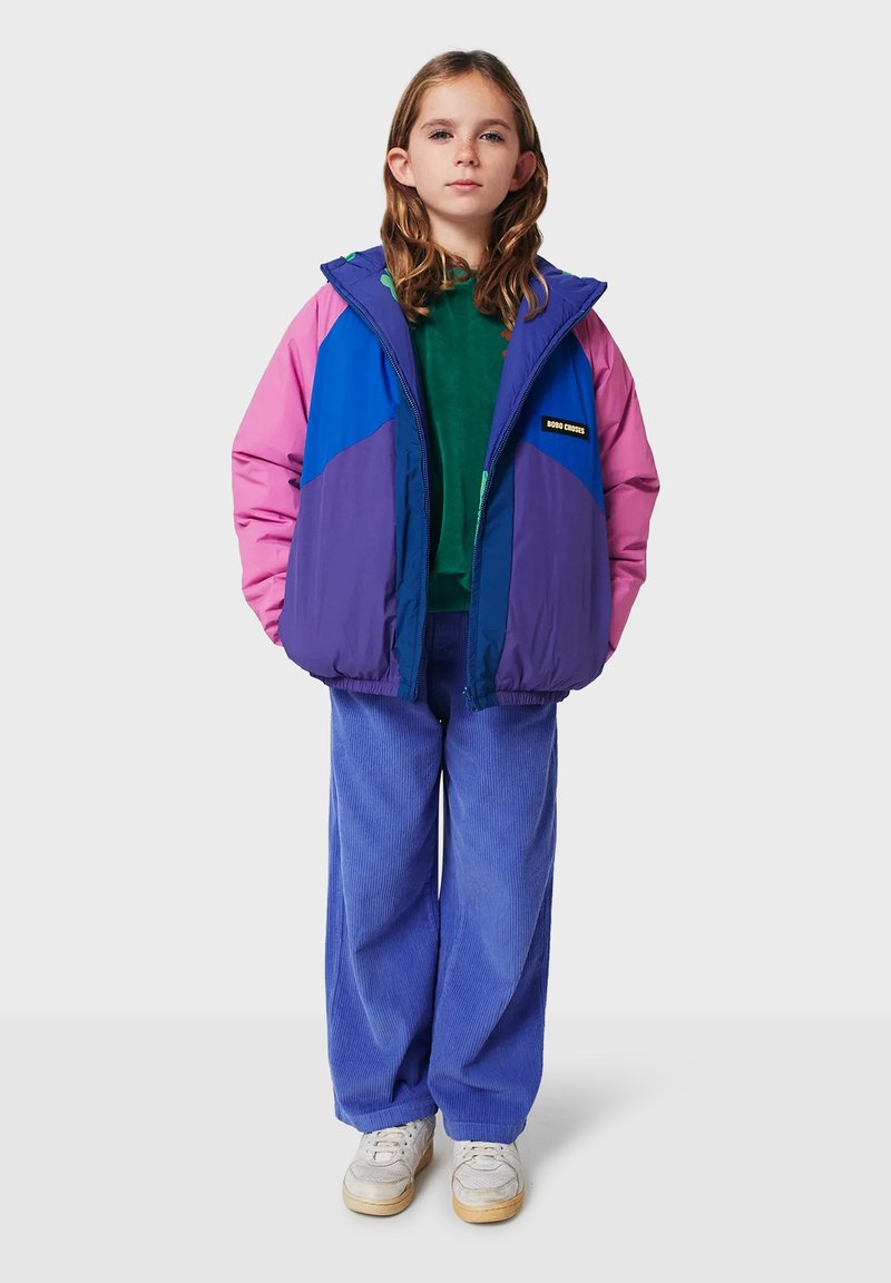 Veste rembourrée colorée avec des panneaux rose, bleu et violet ; dotée d'une capuche et d'un patch logo. Associée à un haut vert, un pantalon en velours côtelé bleu et des baskets blanches.