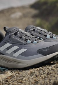 Gros plan sur des chaussures de randonnée Adidas Terrex grises avec des bandes blanches et une étiquette imperméable Rain.RDY sur un terrain rocheux en plein air.
