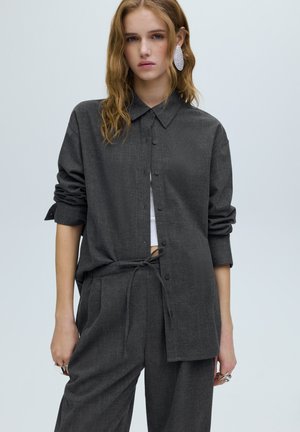 Dunkelgraues Hemd mit Knopfleiste, Kragen und lockerem Schnitt, die Ärmel sind aufgerollt. Kombiniert mit locker sitzenden grauen Hosen, die an der Taille mit einer Schnur gebunden sind.