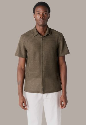 Hombre de pie contra un fondo liso, vistiendo una camisa de manga corta con textura y botones en color verde oliva y pantalones blancos, con los brazos relajados a los lados.