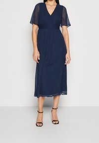 Mörkblå, midjekort klänning med korta, genomskinliga ärmar, en omlott V-ringning och en figursydd midja. Slät textur med flödande kjol.