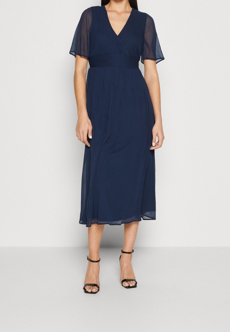 Mörkblå, midjekort klänning med korta, genomskinliga ärmar, en omlott V-ringning och en figursydd midja. Slät textur med flödande kjol.