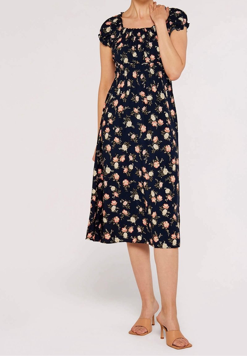 Femme portant une robe midi fleurie bleu marine avec des manches courtes bouffantes et des sandales à talons ouvertes couleur beige, debout devant un fond uni.