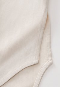 Light beige fabric close-up with a textured finish. Features a curved hem and visible stitching along the edges. Smooth appearance.