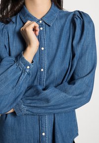 Persona con camisa de mezclilla azul con mangas abullonadas y botones a presión de perla, mano cerca del cuello en un fondo neutro.