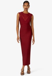 Rotes, eng anliegendes Kleid mit strukturiertem Oberteil, transparenter Detailgestaltung am Halsausschnitt und gerader Silhouette, die bis zur Mitte der Wade reicht. Kombinieren Sie es mit schwarzen Riemchenschuhen.