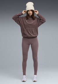 Eine Frau trägt einen mauvefarbenen Cord-Sweatshirt, passende Leggings, eine weiße Mütze und helllila Sneakers, während sie ihren Hut vor einem grauen Hintergrund anpasst.