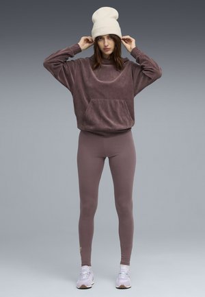 Eine Frau trägt einen mauvefarbenen Cord-Sweatshirt, passende Leggings, eine weiße Mütze und helllila Sneakers, während sie ihren Hut vor einem grauen Hintergrund anpasst.