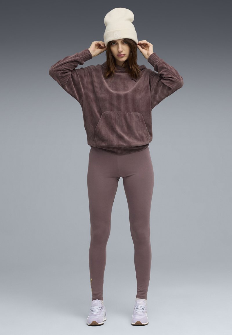 Femme portant un sweat-shirt en corduroy mauve, des leggings assortis, un bonnet blanc et des baskets lilas, ajustant son chapeau sur fond gris.