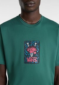 Zelené bavlněné tričko s centrálním grafickým motivem: růžový design mozku orámovaný černým rámem a modrými akcenty, s textem "VANS" na spodní části.