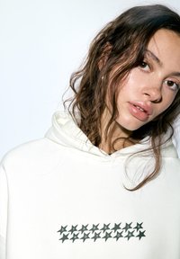 Fehér kapucnis pulóver puha anyagból, elülső részén fekete csillagok grafikájával és a "STARS IN MOTION" felirattal. Állítható kapucnival rendelkezik.