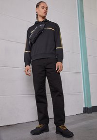 Sweat à capuche noir avec accents dorés, assorti à un pantalon noir. Chaussures noires avec lacets jaunes. Sac bandoulière porté en travers de la poitrine.