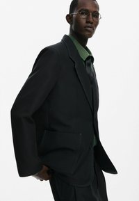 Fekete, szabott blézer kontrasztos varrással, zöld inggel és fekete nyakkendővel párosítva. Szerkezeti kialakítással és dupla zsebbel rendelkezik.