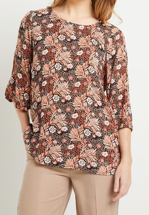 Femme portant un haut ample à motif floral avec des feuilles et des fleurs roses et beiges, associé à un pantalon beige, debout avec une main dans la poche.