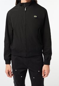 Homme portant une veste noire zippée avec des poignets et une ceinture côtelés, avec un petit logo crocodile vert sur la poitrine.