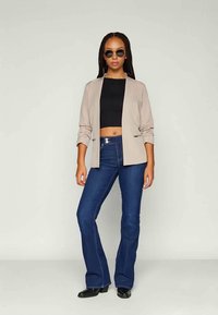 Blazer beige con una vestibilità rilassata, top corto nero e jeans a zampa d'elefante blu scuro. Il modello indossa occhiali da sole scuri, in piedi su uno sfondo chiaro.