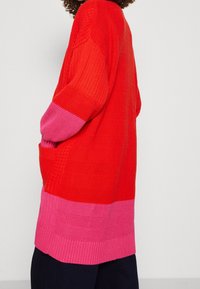 Osoba ubrana w długi, teksturowany sweter typu cardigan z czerwonym górnym i różowym dolnym blokiem kolorów, stojąca na tle jasnego, jednolitego tła.