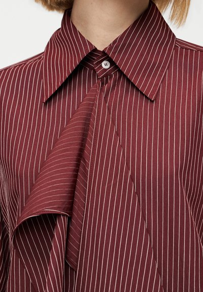 Blouse bordeaux à rayures blanches verticales, avec un col pointu, une fermeture à boutons et un détail de tissu drapé à l'avant.