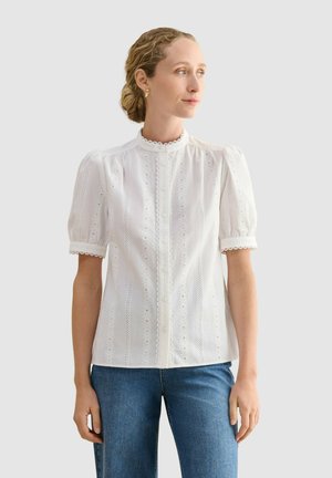 Mujer con el pelo rubio recogido lleva una blusa blanca de encaje calado con mangas abullonadas y vaqueros azules, mirando hacia la derecha.
