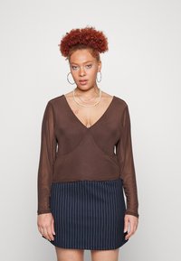 Top castanho de manga longa com decote em V e design cruzado, acompanhado de uma saia mini riscada azul-marinho. O conjunto inclui colares em camadas.