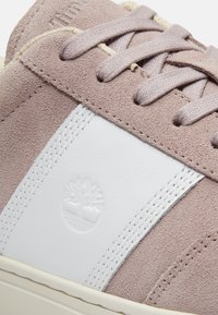 Gros plan d'une basket en daim beige avec un panneau latéral en cuir blanc, un logo d'arbre embossé et des lacets beige assortis.