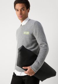 Pilkas vilnonis megztinis su žaliu "FUTURE" logotipu; modelis laiko juodą, ploną, lygaus paviršiaus rankinę prieš neutralią foną.