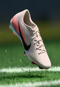 Chuteira de futebol Nike, rosa claro com gradiente de laranja e azul, superfície texturizada, logotipo preto e cravos laranja, em movimento acima da relva.