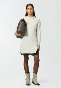 Abito maglione in crema con orlo in pizzo, fessure laterali, abbinato a una borsa marrone scuro. La modella indossa stivali cowboy grigi con dettagli metallici.