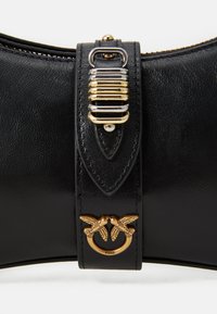 Κοντινό πλάνο από λουρί μαύρης δερμάτινης τσάντας με χρυσό και ασημένιο μεταλλικό κούμπωμα και χρυσό διακοσμητικό κούμπωμα σε σχήμα πουλιού.