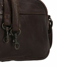 Angolo di una borsa in pelle marrone scuro con due ganci metallici attaccati a cinturini in pelle cuciti, che mostrano texture e dettagli di cucitura.