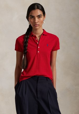 Femeie cu păr lung împletit poartă tricou polo roșu și pantaloni plisati naval, stând pe fundal neutru cu mâinile în buzunare.