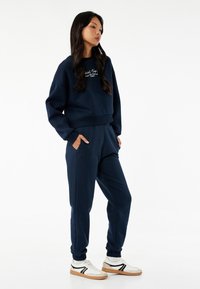 Marineblå cropped sweatshirt med broderet tekst, parret med matchende joggers med sidelommer og elastiske manchetter, båret med hvide sneakers.
