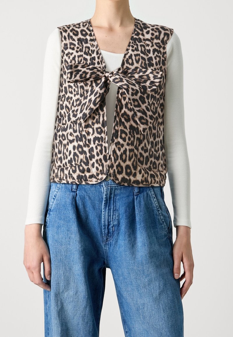 Veste sans manches imprimé léopard avec un grand nœud sur le devant, portée par-dessus un haut blanc à manches longues et un jean bleu ample.