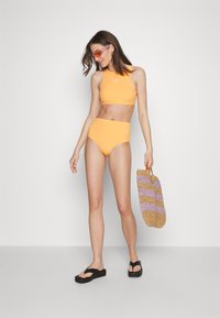 Gelbes Bikini-Set mit einem cropped Top und einer hoch taillierten Hose. Enthält eine geflochtene Tasche in pastellfarbenen Streifen und schwarze Plateausandalen.