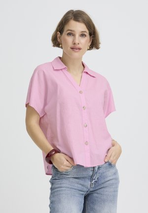 Kobieta z krótkimi brązowymi włosami w różowej koszuli na guziki, niebieskich jeansach, złotych kolczykach kołach i czerwonej bransoletce, z rękami w kieszeniach.