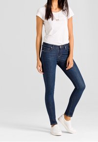 Tmavě modré úzké džíny vyrobené z denimu, nošené s přiléhavým bílým tričkem a bílými nazouváky. Jednoduchý design s jemným prošíváním.