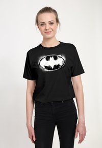 Mladá žena v černém tričku s logem Batmana a černých džínách stojí před čistým bílým pozadím a mírně se usmívá.