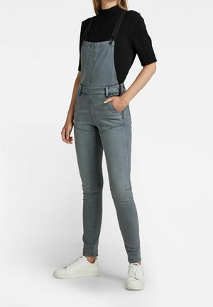 Kobieta ubrana w dopasowane szare dżinsowe ogrodniczki i czarny sweter z krótkim rękawem, z jedną ręką w kieszeni, stojąca na białym tle.