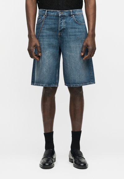 Άνδρας μοντέλο που φορά μπλε τζιν σορτς μέχρι το γόνατο με αλυσίδα, μαύρες κάλτσες και μαύρα δερμάτινα loafers σε λευκό φόντο.