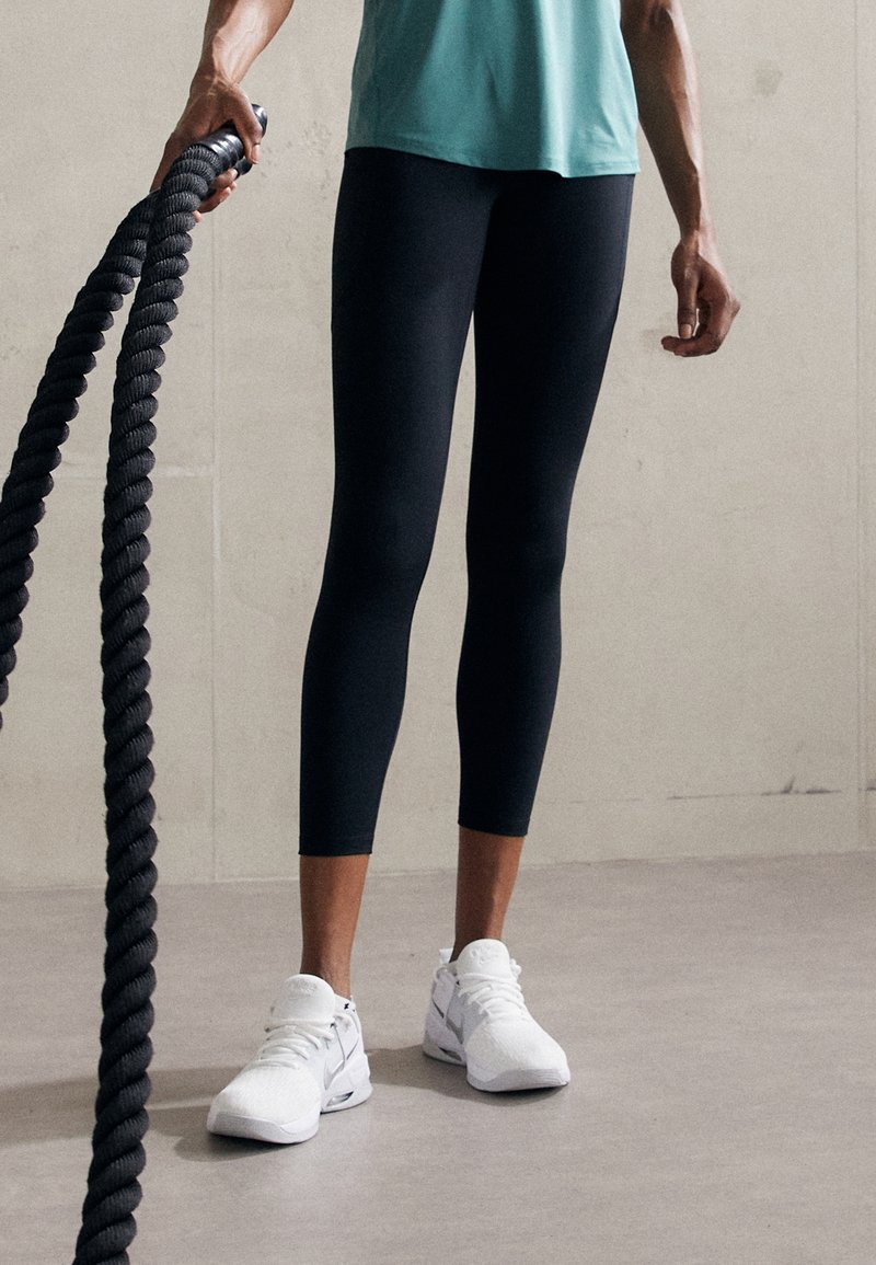 Personne en leggings noirs et sneakers blancs se tenant debout, tenant une épaisse corde de combat noire contre un fond intérieur uni.