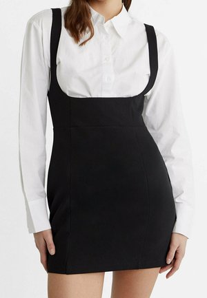 Vrouw die een wit, lang mouws overhemd met knopen draagt onder een nauwsluitende, zwarte mouwloze minijurk met brede bandjes.