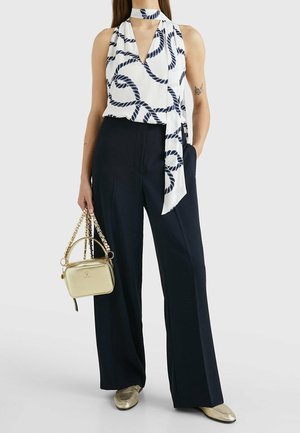 Femme portant un haut blanc sans manches avec un motif de corde marine bleu marine et une écharpe assortie, pantalon large noir, chaussures dorées, tenant un sac à main doré.