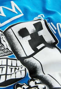 Pikselowany czarno-biały bohater kreskówki trzymający książkę na niebieskim tle materiału z białymi i czarnymi elementami graficznymi.