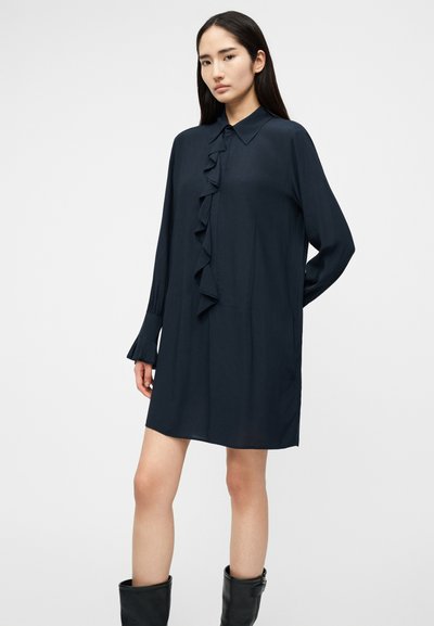 Νεαρή γυναίκα που φοράει ένα μακρυμάνικο σκούρο φόρεμα με βολάν στο μπροστινό μέρος, στέκεται με το ένα χέρι πίσω από την πλάτη της και μαύρες μπότες μέχρι τη μέση της γάμπας.